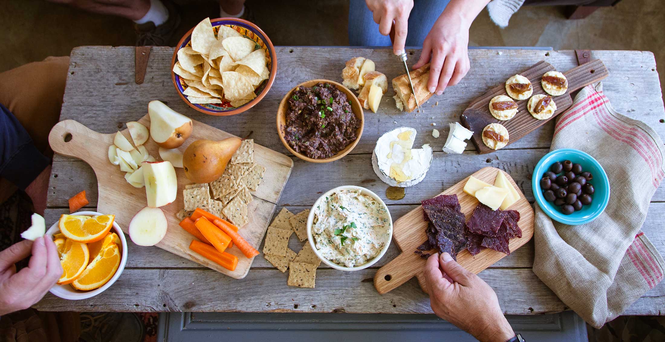 Hands reach across a wooden table to pick up bites of Patagonia Provisions organic snacks and appetizers in bowls and on cutting boards for a shared meal